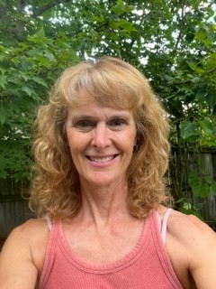 Woman with with shoulder length blond hair in a pink tank top with green leaves in the background